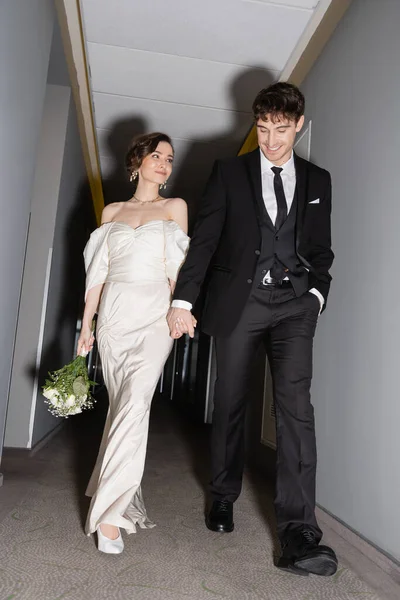 Low angle view of cheerful groom in black suit posing with hand in pocket and holding hand of bride in white wedding dress carrying bridal bouquet while walking together in hall of modern hotel — Stock Photo