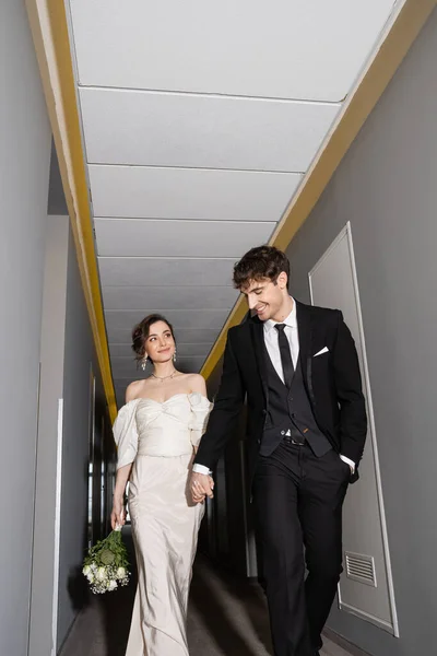 Низький кут зору веселого нареченого в чорному костюмі, тримаючи руку чудової нареченої в білій весільній сукні, що несе весільний букет, ходячи разом у залі сучасного готелю — Stock Photo