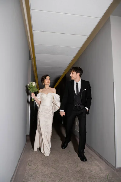 Full length of happy groom in black suit posing while holding hands with bride in white dress carrying bridal bouquet while walking together in corridor of modern hotel — Stock Photo