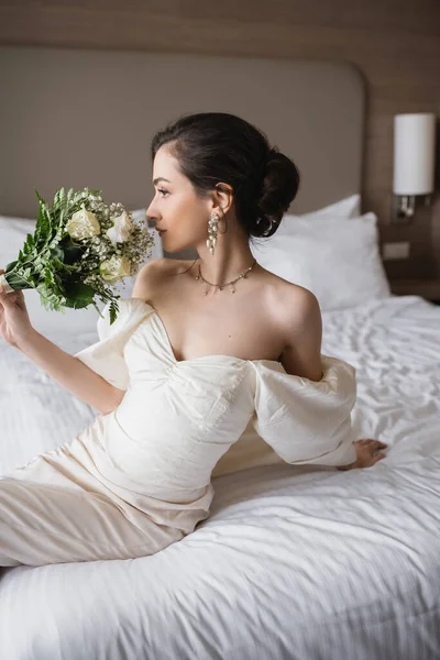Young bride in white dress and luxurious jewelry sitting on bed and smelling bridal bouquet with flowers while looking away in modern bedroom in hotel room on wedding day — Stock Photo