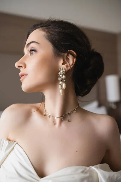 Портрет молодой невесты в белом платье и роскошные украшения с жемчужными серьгами и ожерельем, глядя в современную спальню в гостиничном номере в день свадьбы — стоковое фото