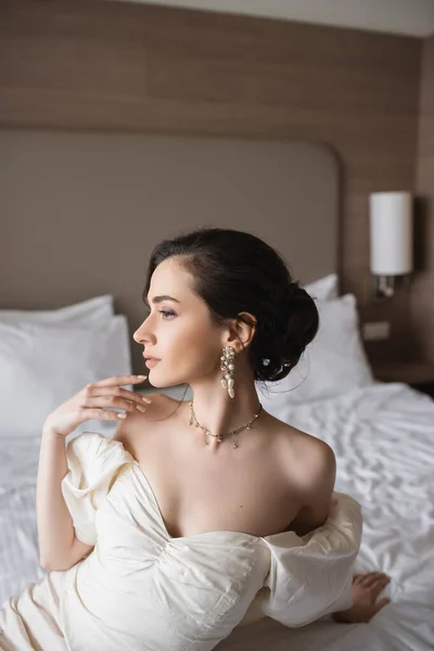 Vista lateral de la encantadora novia joven en vestido blanco y joyas de lujo con pendientes de perlas y collar mirando hacia otro lado en el dormitorio moderno en la habitación de hotel el día de la boda - foto de stock