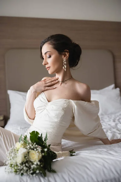 Encantadora novia joven en vestido blanco y joyas de lujo sentado en la cama junto al ramo de novia con flores en el dormitorio moderno de la habitación de hotel en el día de la boda - foto de stock