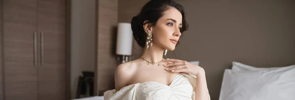 Alluring bride in white dress and luxurious jewelry with pearl earrings and necklace touching chest while sitting in modern bedroom in hotel room on wedding day, banner — Stock Photo