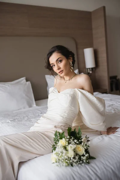 Stunning young bride in white dress and luxurious jewelry sitting on bed next to bridal bouquet with flowers and looking at camera in modern bedroom in hotel room on wedding day — Stock Photo