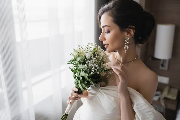Vista laterale di elegante giovane sposa in abito bianco e gioielli di lusso con orecchini di perle che tengono bouquet da sposa in camera d'albergo moderno il giorno del matrimonio — Foto stock