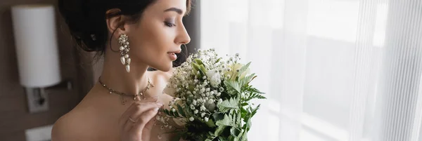 Seitenansicht der eleganten jungen Braut mit luxuriösem Schmuck, in Perlenohrringen und Halskette mit Brautstrauß im modernen Hotelzimmer am Hochzeitstag, Banner — Stockfoto
