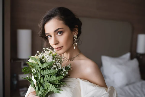 Bezaubernde junge Braut in weißem Kleid und luxuriösem Schmuck hält Brautstrauß mit Blumen in der Hand und blickt in die Kamera im modernen Schlafzimmer im Hotelzimmer am Hochzeitstag — Stockfoto