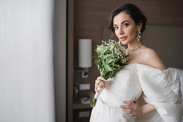 Encantadora jovem mulher em vestido de noiva branco e jóias de luxo segurando buquê de noiva com flores e posando enquanto olha para a câmera no quarto moderno do quarto de hotel — Fotografia de Stock