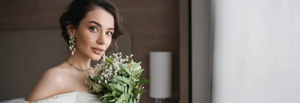 Чарівна молода жінка в білій весільній сукні і розкішні прикраси, що тримають весільний букет з квітами і дивляться на камеру в сучасній спальні в готельному номері, банер — стокове фото