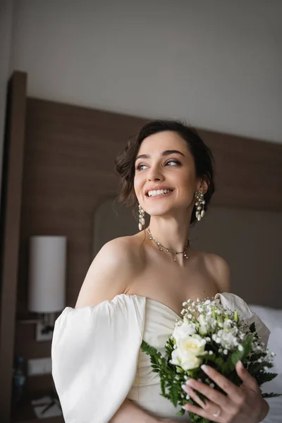 Cheerful young woman in white wedding dress and luxurious jewelry holding bridal bouquet with flowers and looking away in modern bedroom in hotel room — Stock Photo