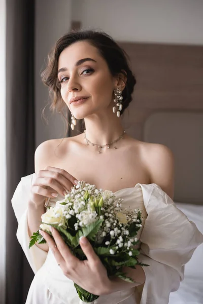 Enchanteresse jeune femme en robe de mariée et bijoux de luxe tenant bouquet nuptiale avec des fleurs et regardant la caméra dans la chambre moderne dans la chambre d'hôtel le jour du mariage — Photo de stock
