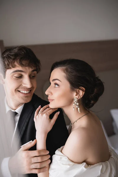Hübsche junge Braut in Schmuck und Brautkleid schaut fröhlichen Bräutigam in schwarzer Festkleidung an, während sie nach der Zeremonie im modernen Hotelzimmer zusammensteht — Stockfoto