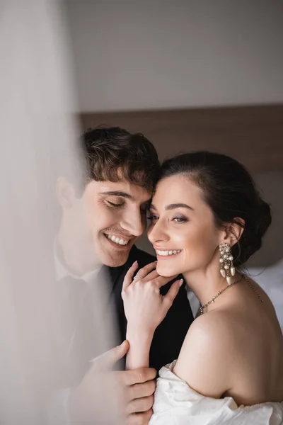 Pretty young bride in jewelry and wedding dress hugging shoulder of cheerful groom in classic formal wear while standing together in modern hotel room after ceremony — Stock Photo