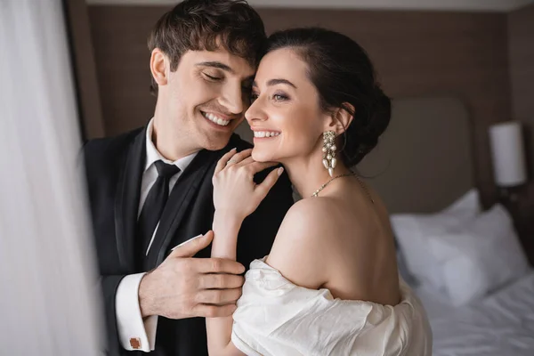 Joyful young bride in jewelry and wedding dress hugging shoulder of cheerful groom in classic formal wear while standing together in modern hotel room after ceremony — Stock Photo