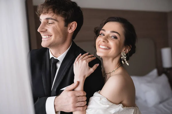 Счастливая невеста в элегантных украшениях и свадебном платье обнимает плечо счастливого жениха в классической формальной одежде, стоя вместе в современном гостиничном номере после церемонии — стоковое фото