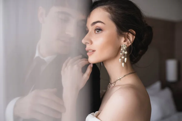 Stunning bride in elegant jewelry and wedding dress hugging shoulder of groom in classic formal wear while standing together behind white tulle in modern hotel room after ceremony — Stock Photo