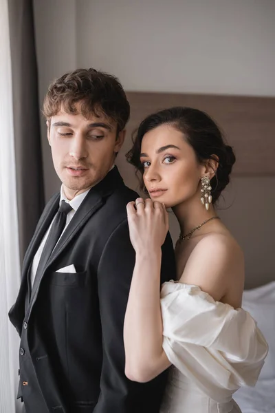 Bride with brunette hair, in elegant jewelry and white dress looking at camera and hugging shoulder of groom in classic formal wear with tie in modern hotel room after wedding ceremony — Stock Photo