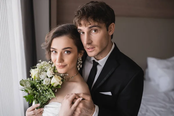 Pretty young bride in jewelry, white dress with bridal bouquet holding hands with groom in classic formal wear while standing together in modern hotel room after wedding ceremony — Stock Photo