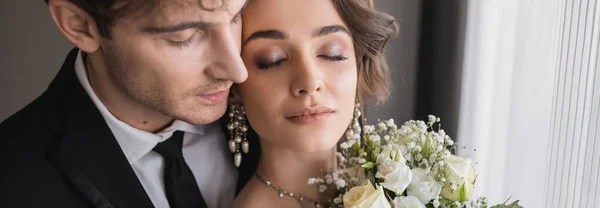 Pretty bride with closed eyes, in jewelry with pearls and bridal bouquet standing next to groom in classic formal wear with tie in modern hotel room after wedding ceremony, banner — Stock Photo