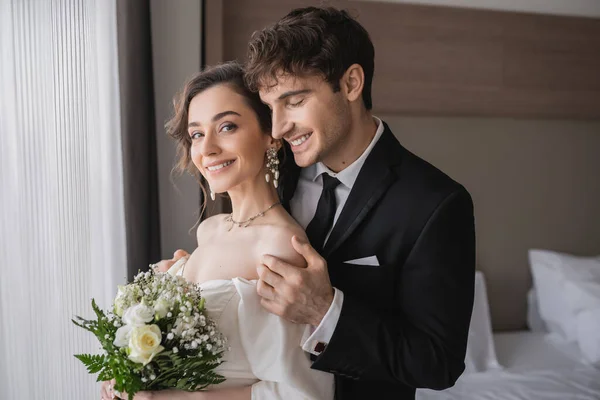 Glücklicher Bräutigam in klassischer formaler Kleidung, der die Schulter einer eleganten jungen Braut in Schmuck berührt, weißes Kleid mit Brautstrauß, während er nach der Trauung im modernen Hotelzimmer zusammensteht — Stockfoto