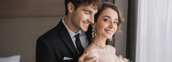 Happy groom in classic formal wear touching shoulder of elegant and cheerful young bride in jewelry with pearls while standing together in modern hotel room after wedding ceremony, banner — Stock Photo