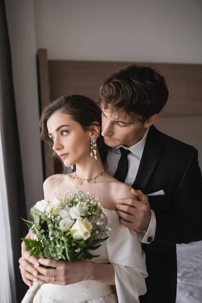 Tender groom in classic formal wear touching shoulder of elegant young bride in jewelry, white dress with bridal bouquet while standing together in modern hotel room after wedding ceremony — Stock Photo