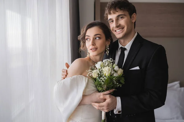 Fröhlicher Bräutigam in klassisch-formaler Kleidung umarmt elegante Braut in Schmuck, weißes Kleid mit Brautstrauß beim Zusammenstehen im modernen Hotelzimmer nach der Trauung — Stockfoto