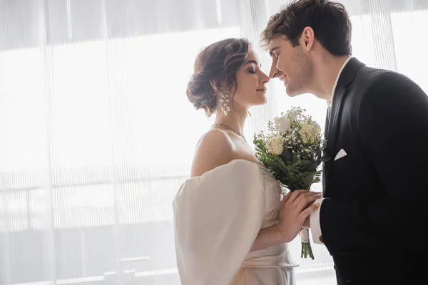 Side view of young bride in jewelry, white dress with bridal bouquet and groom in classic formal wear standing together with closed eyes in modern hotel room after wedding ceremony, happy couple — Stock Photo