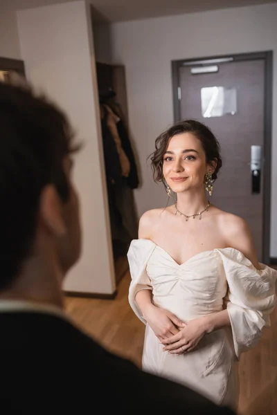 Pretty young bride in elegant jewelry and white dress looking at blurred groom while standing near entrance door to modern hotel room after wedding ceremony, happy couple — Stock Photo