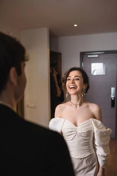 Pretty young bride in elegant jewelry and white dress laughing and looking at blurred groom while standing near entrance door to modern hotel room after wedding ceremony, happy couple — Stock Photo