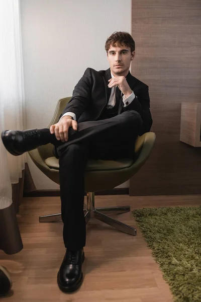 Junger Mann in schwarzer Festkleidung mit Krawatte und klassischen Schuhen sitzt auf bequemen Sesseln und blickt am Hochzeitstag in die Kamera, posiert in modernem Hotelzimmer, gut aussehender Bräutigam — Stockfoto