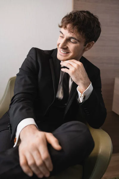 Hombre alegre en ropa formal negro con corbata sentado en un sillón cómodo y mirando hacia otro lado en el día de la boda, descansando en la habitación de hotel moderna, novio guapo — Stock Photo