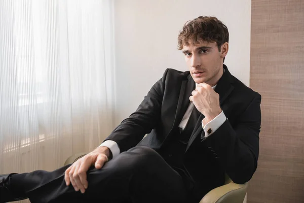Hombre de éxito en ropa formal negro con corbata sentado en un sillón cómodo y mirando a la cámara en el día de la boda, descansando en la habitación de hotel moderna, novio guapo - foto de stock