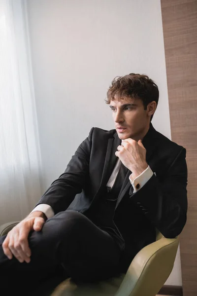 Schöner Mann in schwarzer Festkleidung mit Krawatte sitzt mit der Hand am Kinn auf einem bequemen Sessel und schaut am Hochzeitstag weg, posiert in einem modernen Hotelzimmer — Stockfoto