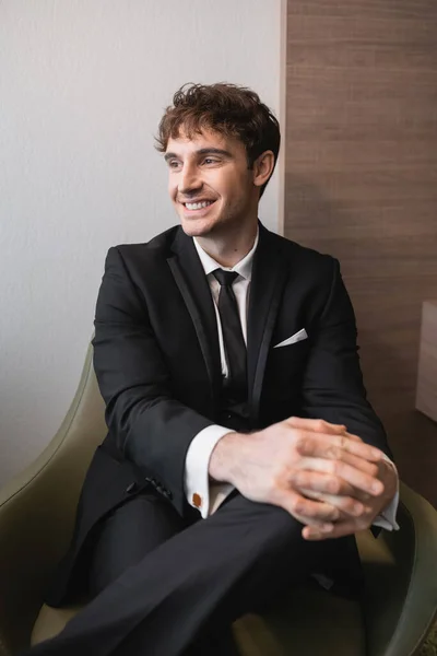 Pleased man in black formal wear with tie sitting on comfortable armchair and looking away on wedding day, resting in modern hotel room before marriage, handsome groom — Stock Photo