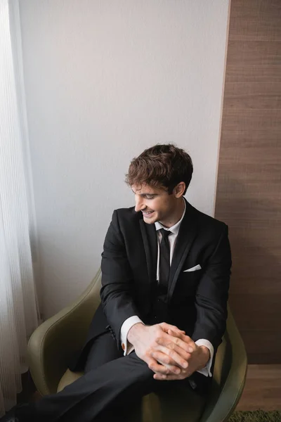 Счастливый человек в черном костюме с галстуком улыбается и сидит с сжатыми руками на удобном кресле и смотрит в сторону в день свадьбы, отдыхает в современном гостиничном номере, красивый жених — стоковое фото