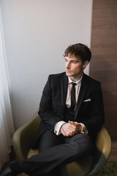 Молодий чоловік в чорному костюмі з краваткою, сидячи з затишними руками на зручному кріслі і дивлячись в день весілля, думаючи в сучасній кімнаті готелю перед шлюбом, гарний вигляд нареченого — стокове фото
