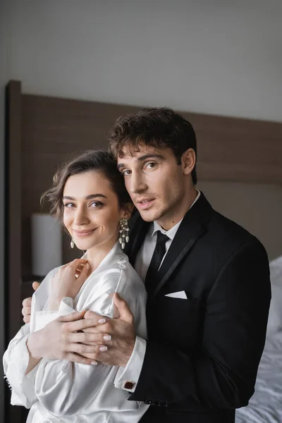Bräutigam in klassischer formeller Kleidung umarmt glückliche junge Braut in Schmuck und weißem Kleid, während sie zusammen in einem modernen Hotelzimmer während ihrer Flitterwochen nach der Hochzeit stehen — Stockfoto