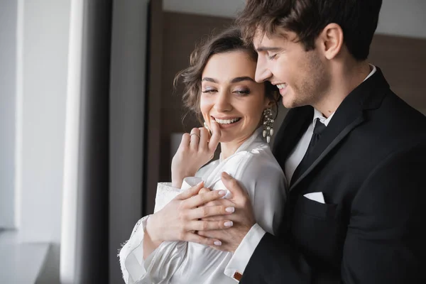 Noivo em terno preto clássico abraçando a noiva jovem feliz em jóias e vestido branco enquanto estão juntos no quarto de hotel moderno durante sua lua de mel no dia do casamento, recém-casados alegres — Fotografia de Stock