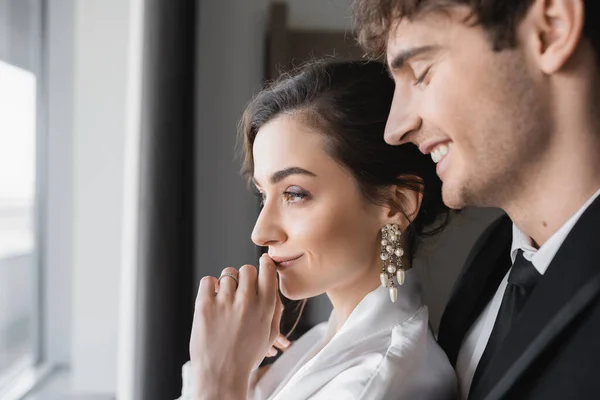 Веселый жених в классическом черном костюме, улыбающийся рядом с молодой невестой в ювелирном и белом свадебном платье, стоя вместе в современном гостиничном номере во время медового месяца, счастливые молодожены, рука у губ — стоковое фото