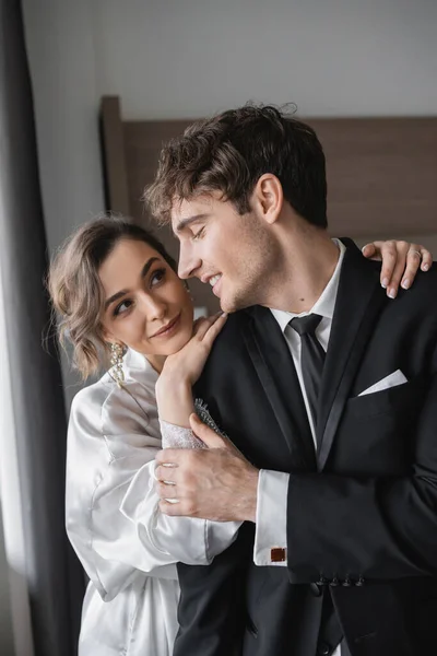 Pleased bride in jewelry and white silk robe leaning on shoulder of joyful groom in classic black suit while standing together in modern hotel room during honeymoon, newlyweds — Stock Photo