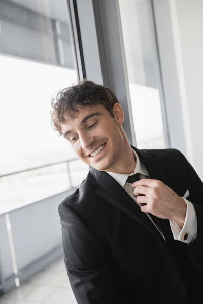Happy man in classic formal wear adjusting black tie while doing preparations and standing in modern hotel room near window, groom on wedding day, special occasion — Stock Photo