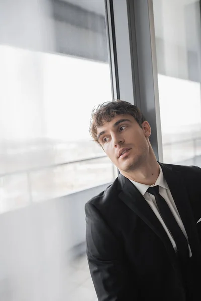 Thoughtful man in classic formal wear with black tie and white shirt standing in modern hotel room and looking at window, groom on wedding day, special occasion — Stock Photo
