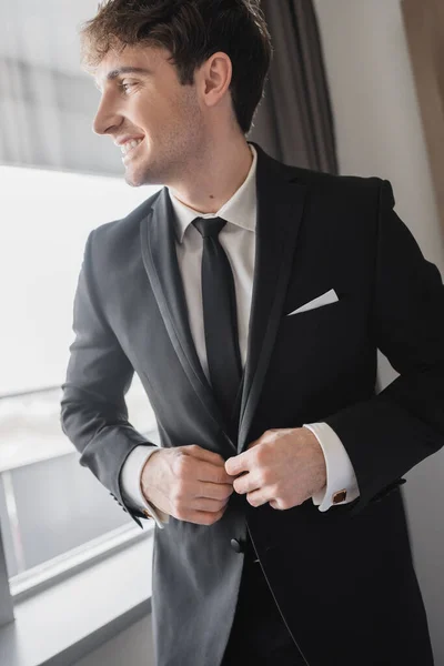 Happy man in classic formal wear with black tie and white shirt buttoning blazer and standing in modern hotel room near window, groom on wedding day, special occasion — Stock Photo