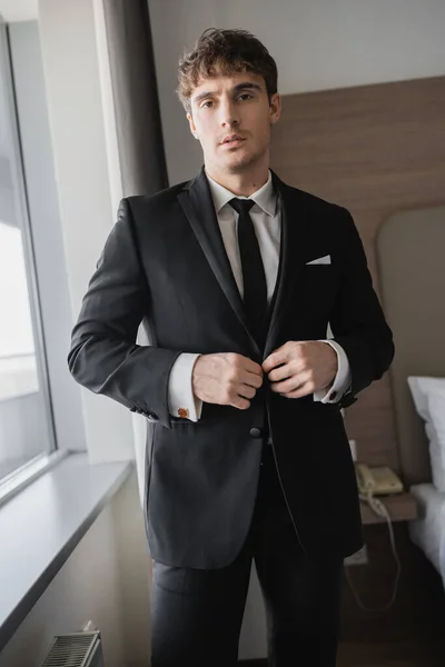Красивый мужчина в стильной формальной одежде с черным галстуком и белой рубашкой застегивающий пиджак и стоя в современном гостиничном номере рядом с окном, жених в день свадьбы, особый случай — стоковое фото