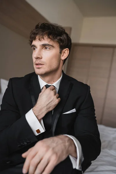 Красивий чоловік у стильному формальному одязі з білою сорочкою, що регулює чорну краватку, дивлячись у сучасну готельну кімнату, наречений у день весілля, особливий випадок — стокове фото