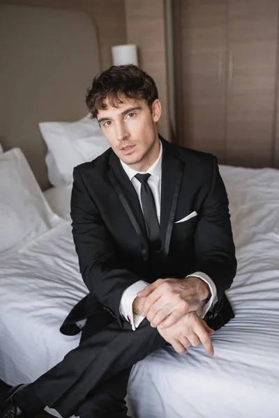Schöner Mann in eleganter formaler Kleidung mit schwarzer Krawatte und weißem Hemd sitzt auf dem Bett im modernen Hotelzimmer und blickt in die Kamera, Bräutigam am Hochzeitstag, besonderer Anlass — Stockfoto