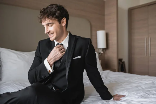 Fröhlicher Mann in eleganter formaler Kleidung mit weißem Hemd und schwarzer Krawatte, während er wegschaut und im modernen Hotelzimmer auf dem Bett sitzt, Bräutigam am Hochzeitstag, besonderer Anlass — Stockfoto