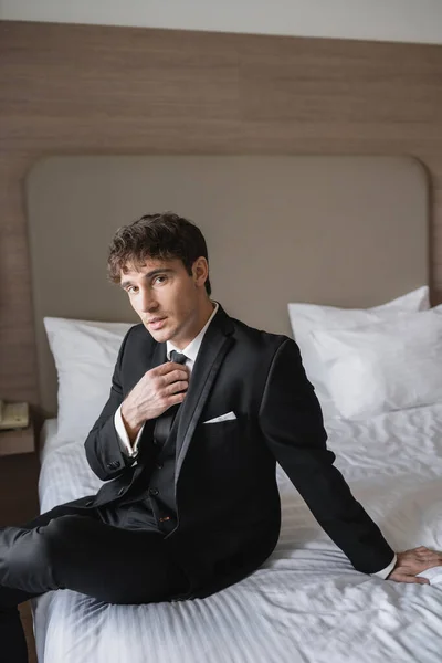 Hombre guapo en ropa formal con clase con camisa blanca que ajusta la corbata negra mientras mira a la cámara y se sienta en la cama en la habitación de hotel moderna, novio en el día de la boda, ocasión especial - foto de stock
